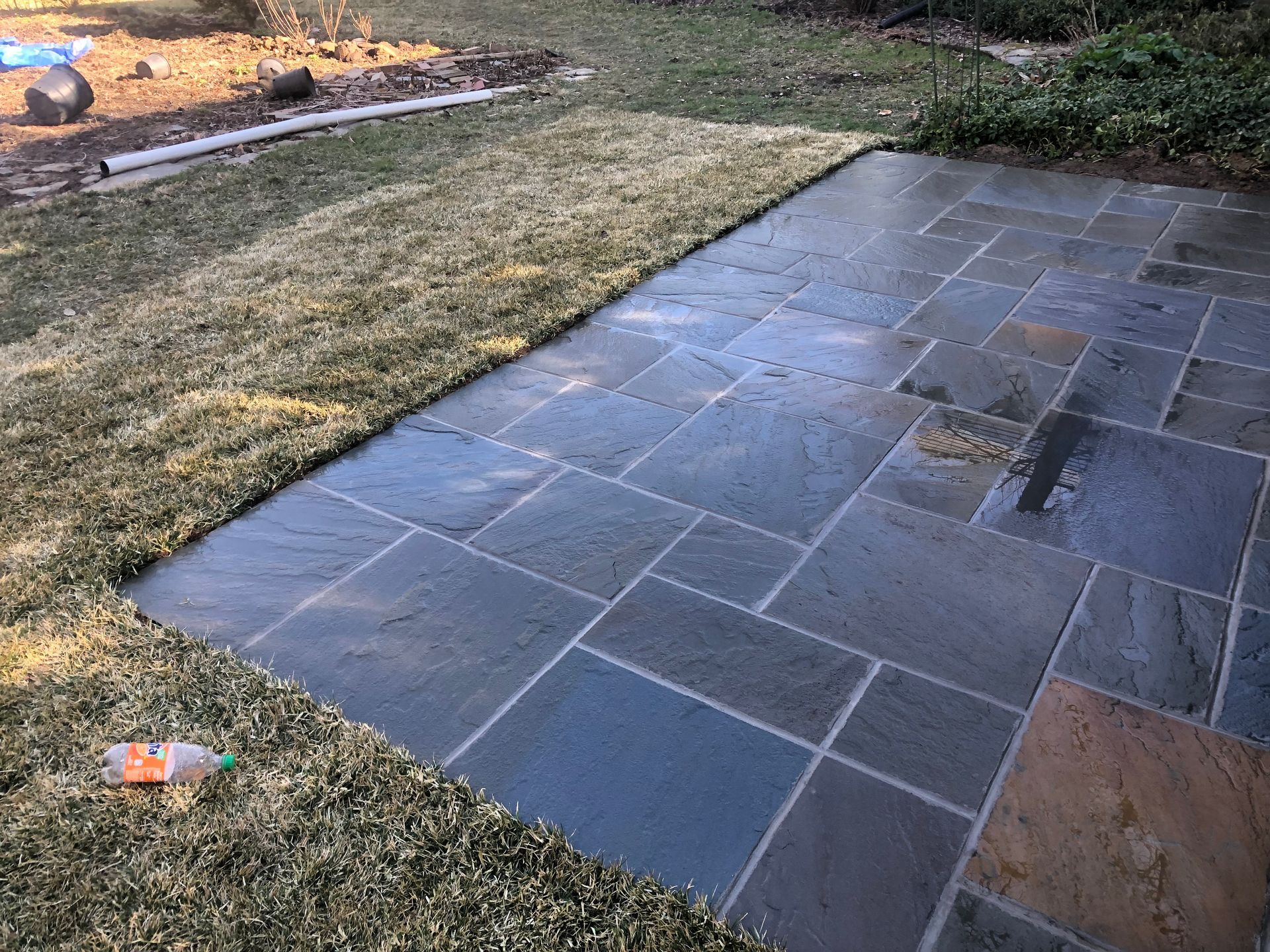 Dark stone patio beside a patchy lawn, with a landscaped garden bed in the background.