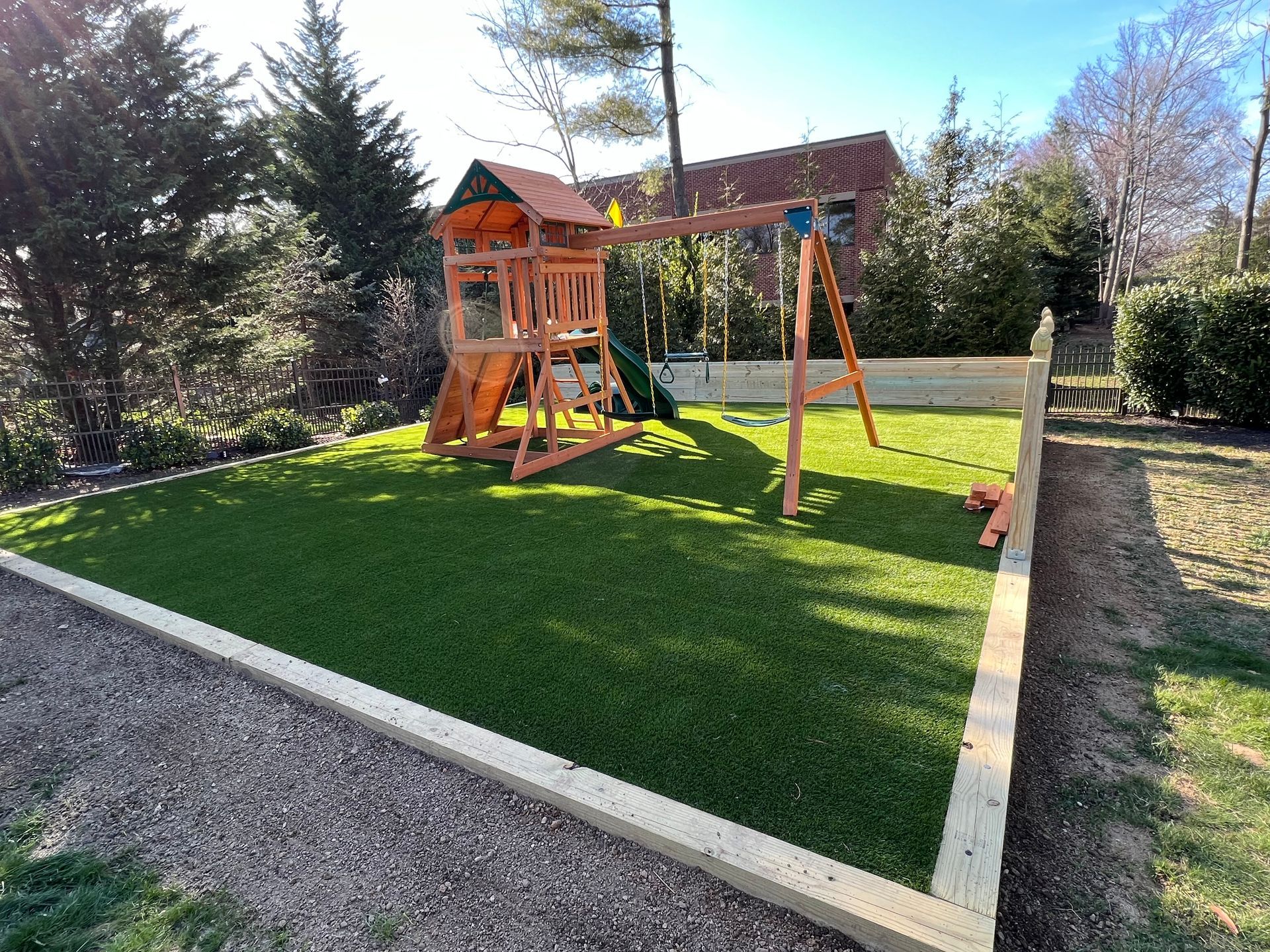 Backyard playset on a green lawn with a swing set and wooden fort, bordered by gravel and stone edging