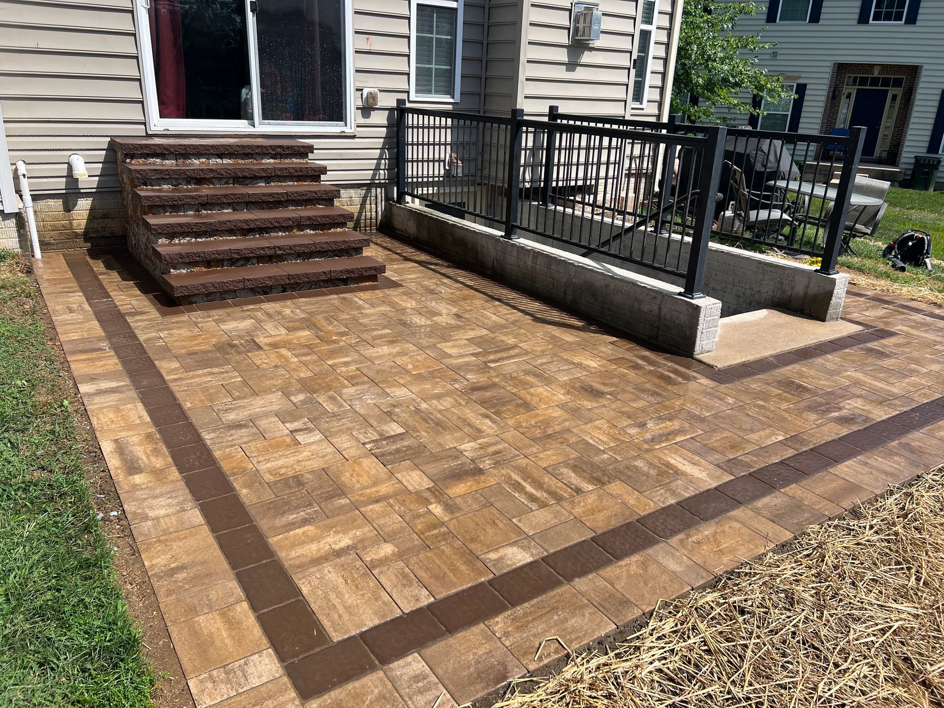 Brick patio with steps and black railing beside a house, bordered by grass and mulch.