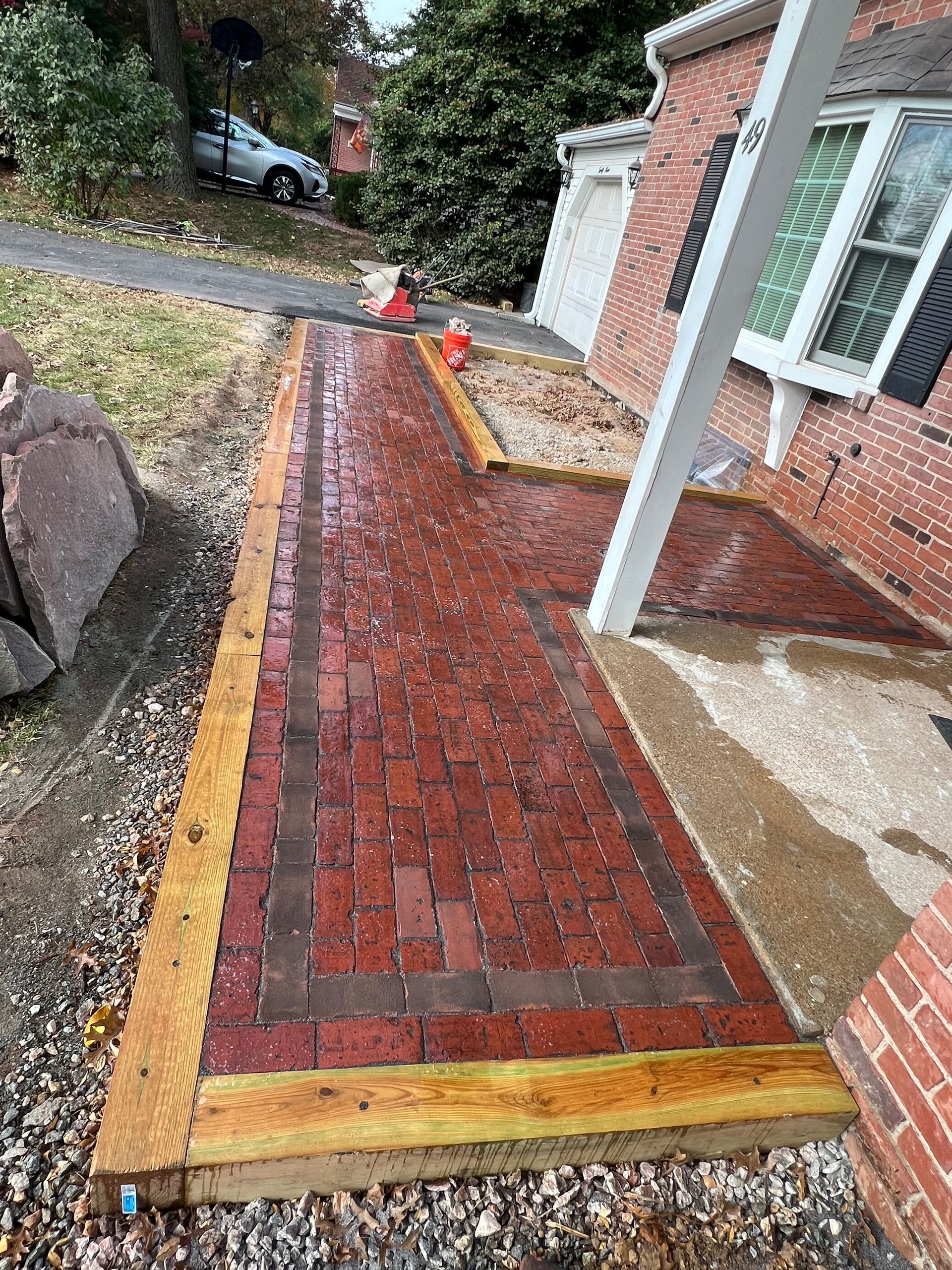 Wet brick walkway bordered by stone and wood, leading to a brick house with white trim.