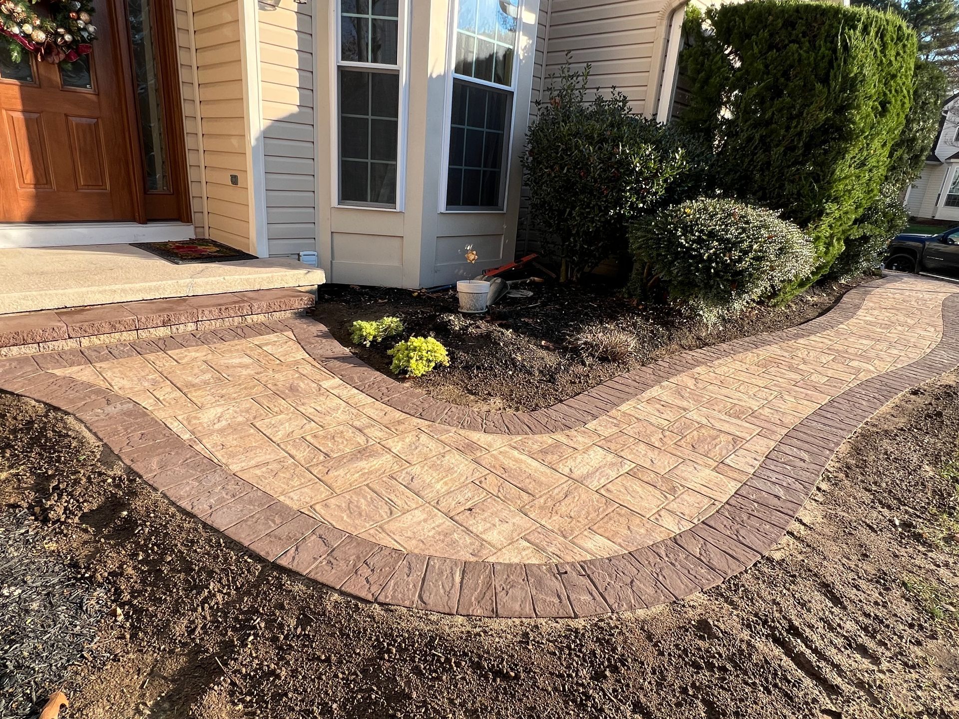 Curved brick walkway and mulch border beside a house entrance with shrubs and a bay window