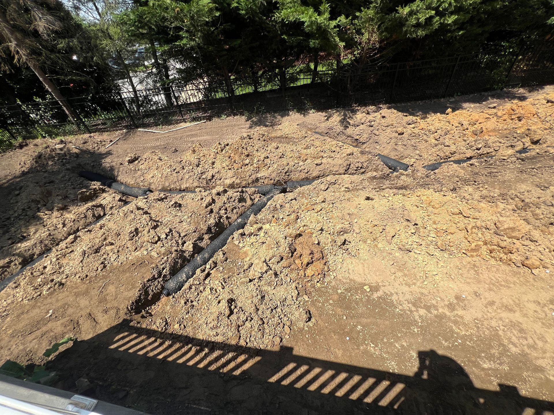 Excavated dirt lot with trenches and a metal grate in the foreground, trees along the edge.