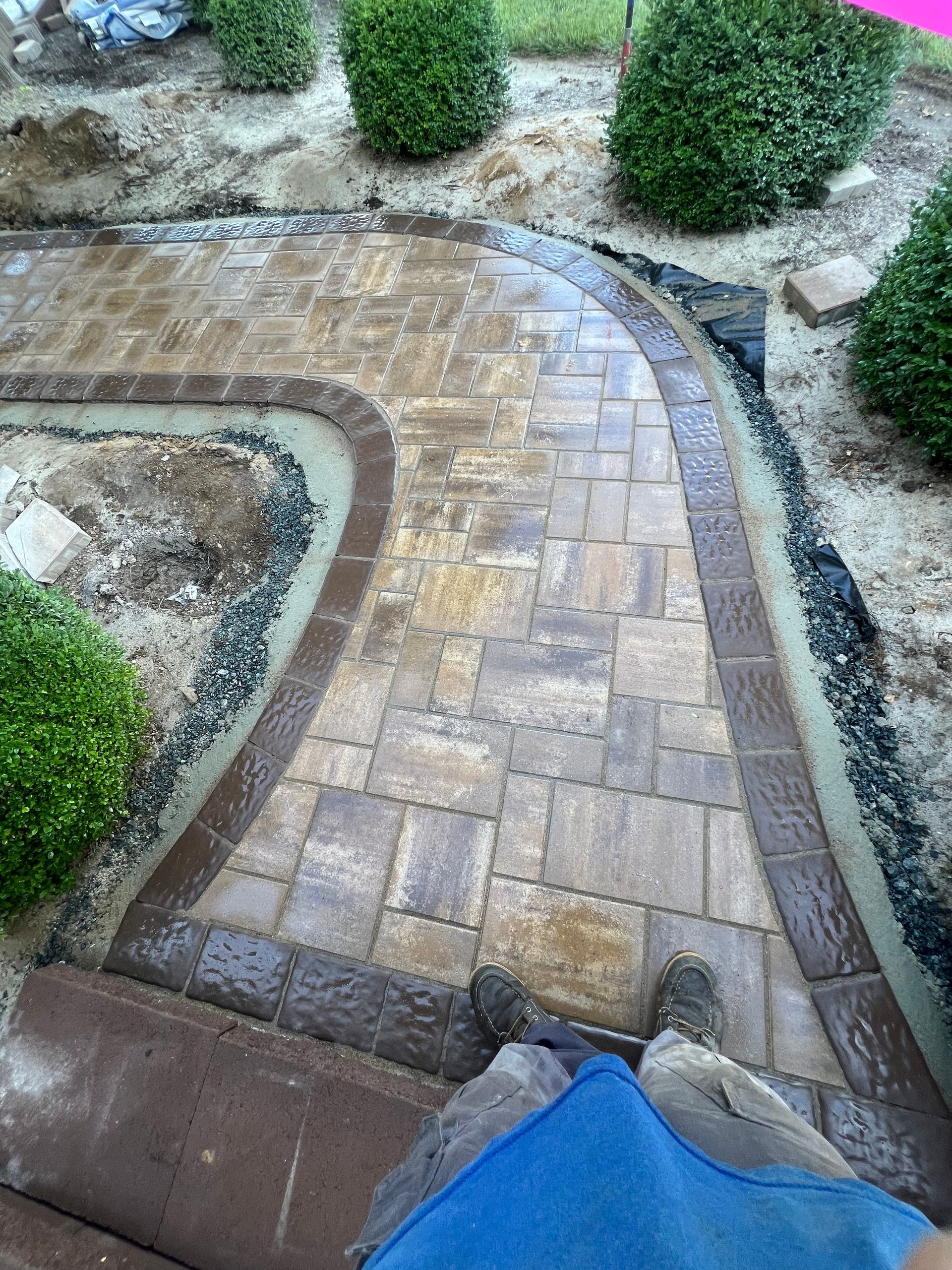 Curved brick walkway through a garden with shrubs and a blue tarp at the bottom