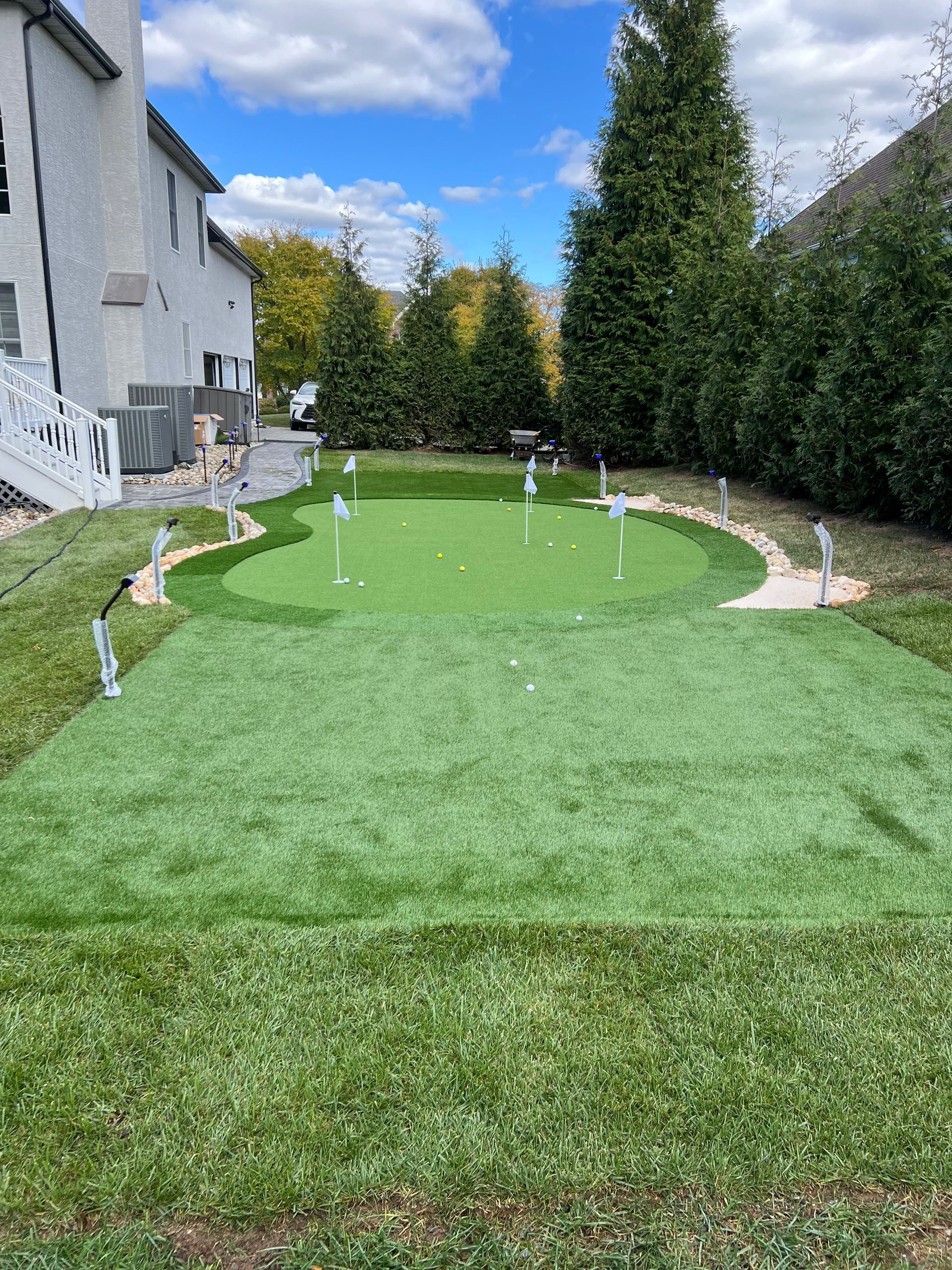Backyard putting green with short artificial turf, golf flags, and tall hedges beside a house