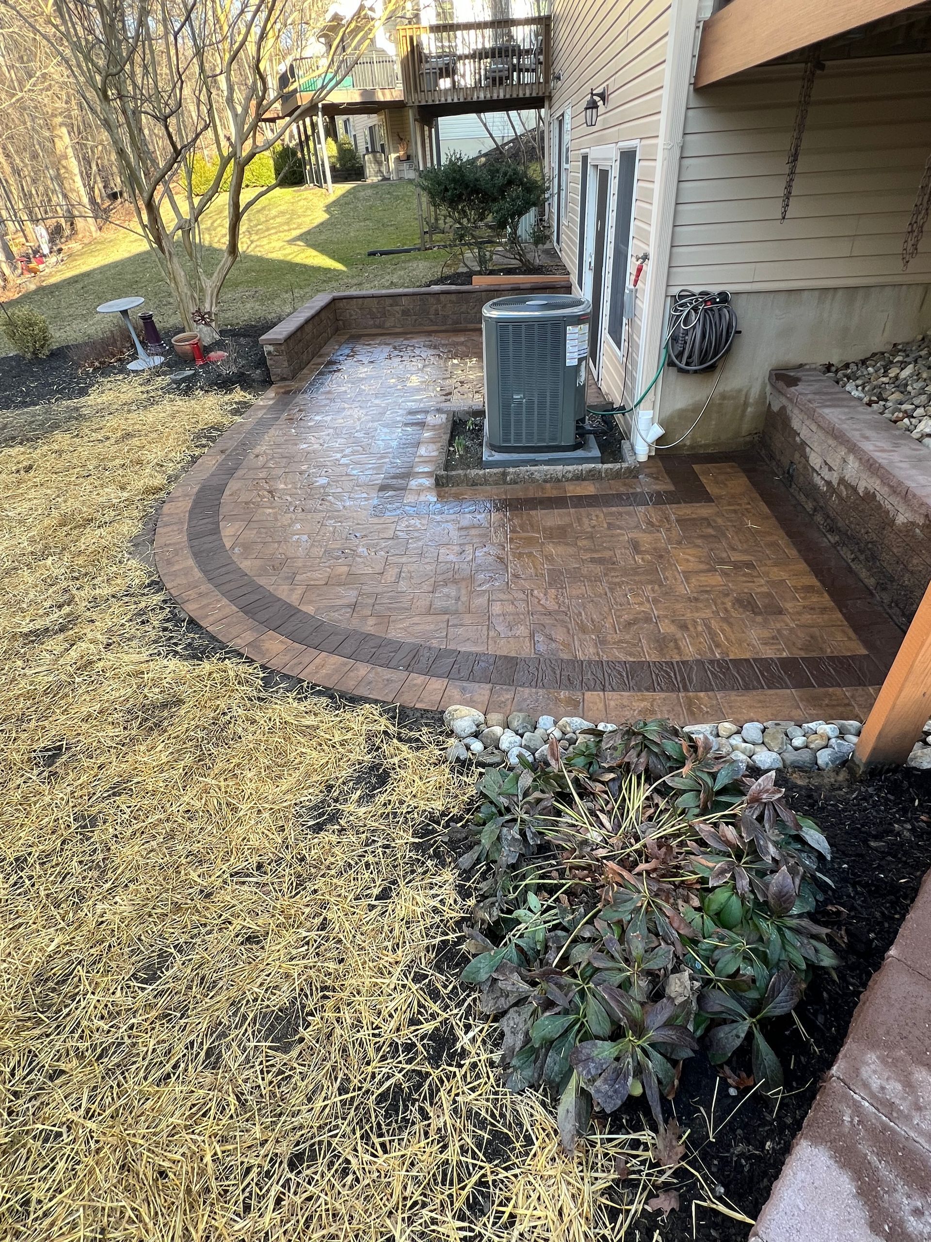 Curved brick patio beside a house, with an outdoor HVAC unit and a grassy yard covered in fallen leaves.