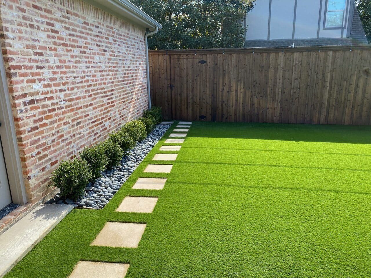 Backyard with green lawn, brick house wall, wooden fence, and stepping stones along the edge