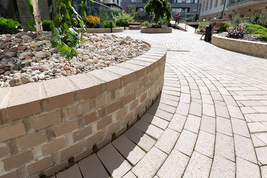 Curved brick retaining wall beside a light stone walkway in an outdoor plaza with plants.