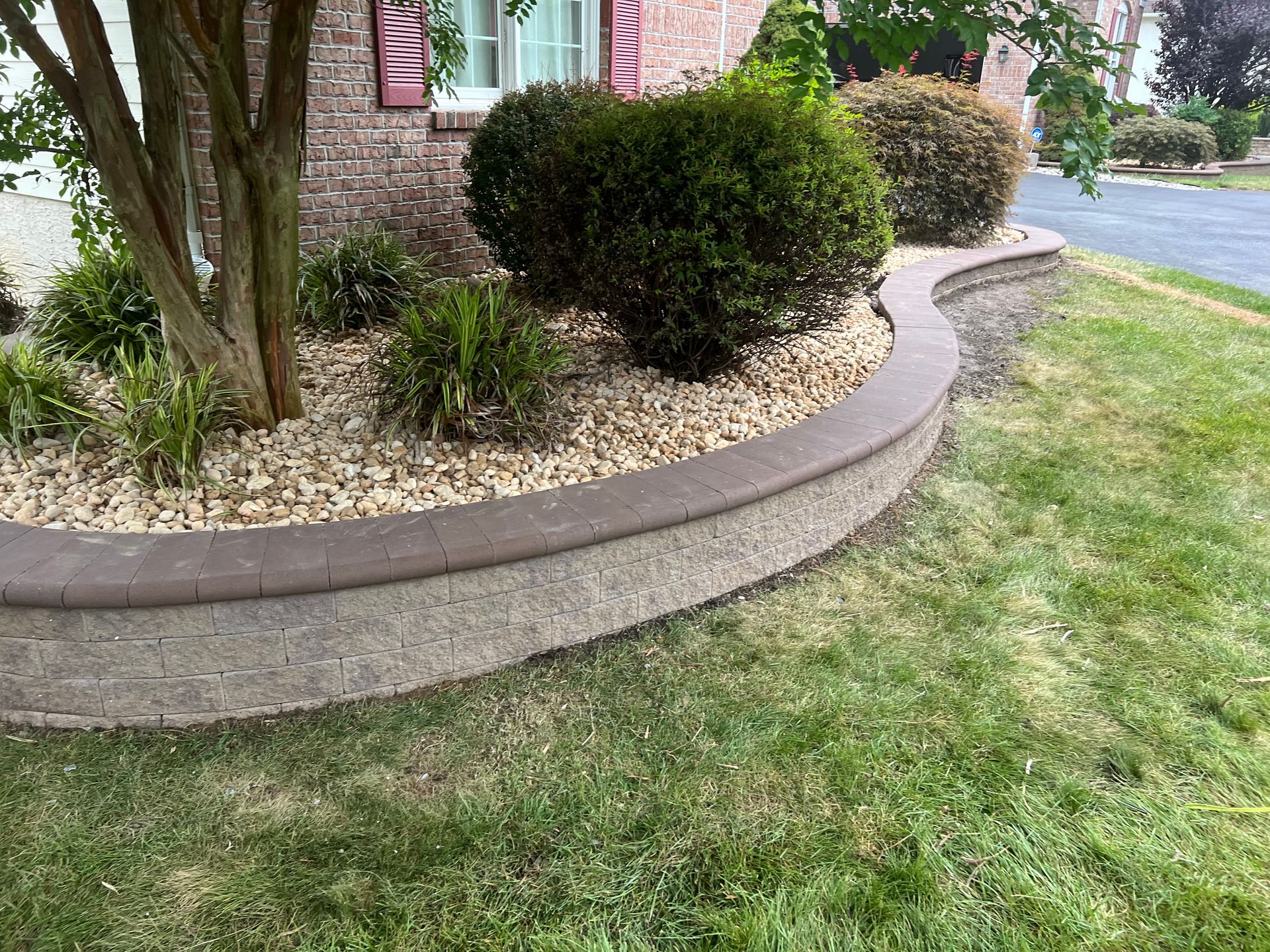 Curved stone garden border with shrubs and mulch beside a grassy lawn and house exterior