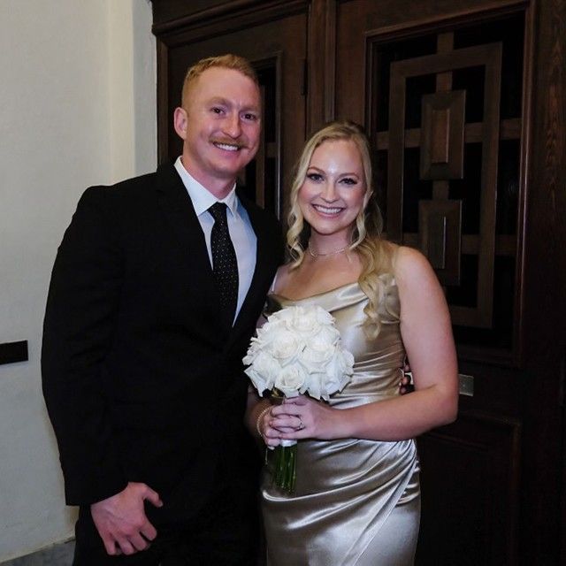 A man in a suit and tie stands next to a woman in a dress holding a bouquet of white flowers