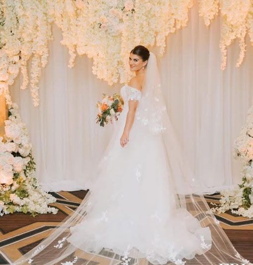A bride in a wedding dress and veil is standing in front of a wall of white flowers.