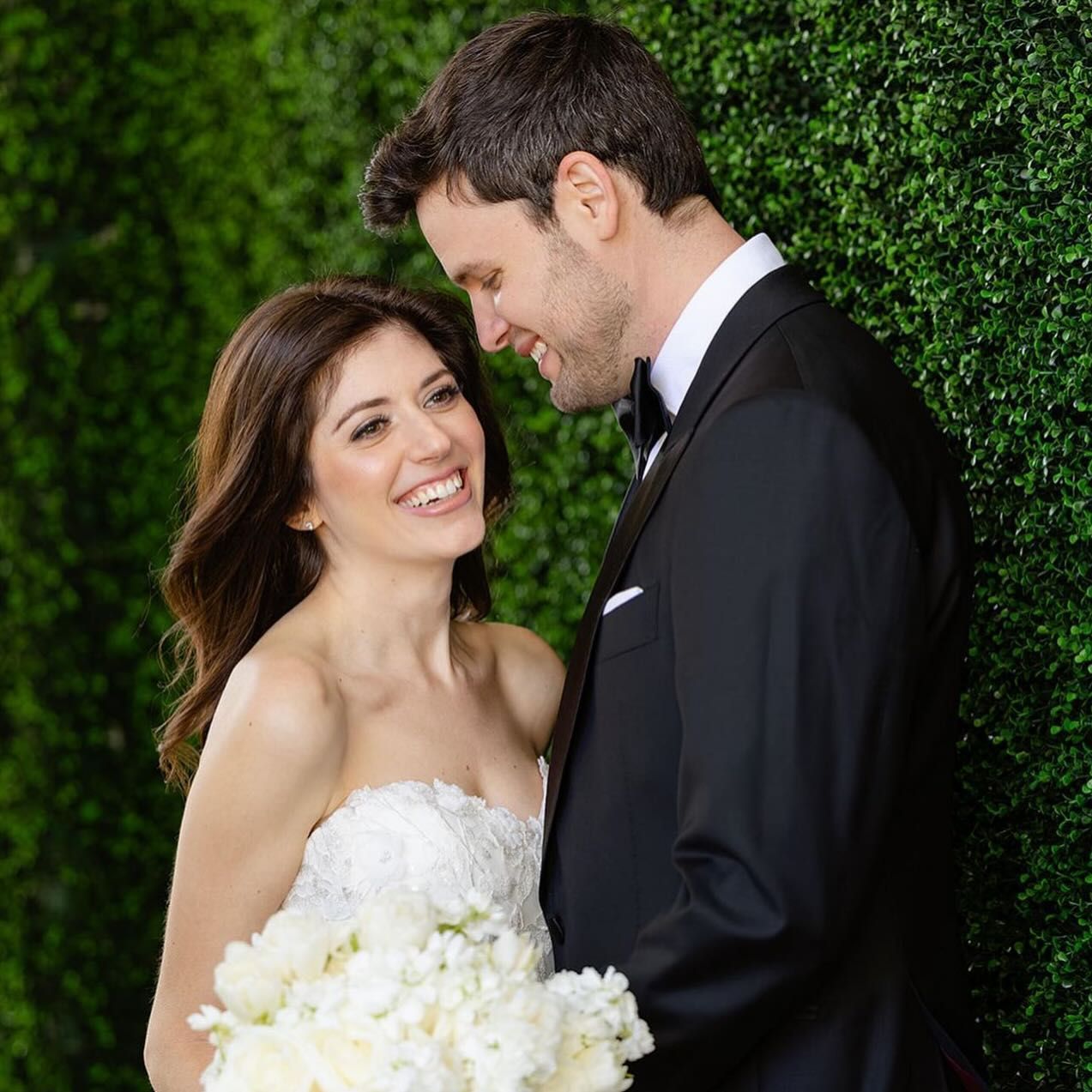 A bride and groom are looking at each other in front of a green hedge.