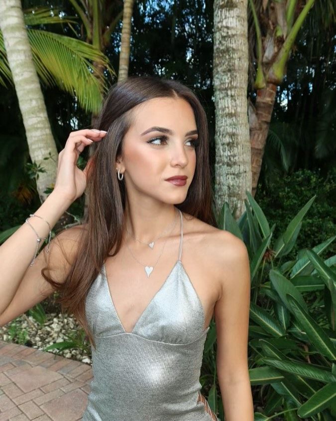 A woman in a silver top is standing in front of palm trees.
