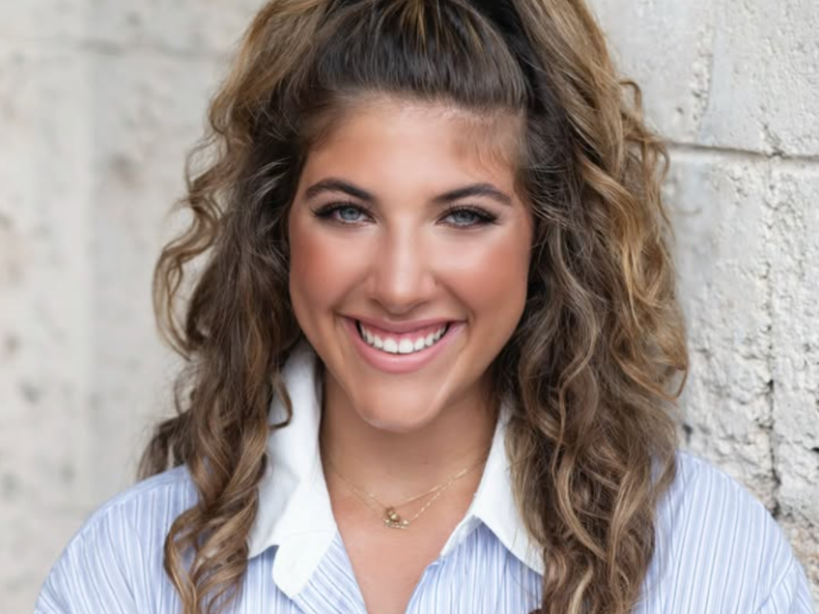 A woman with very long brown hair is standing in front of a brown background.