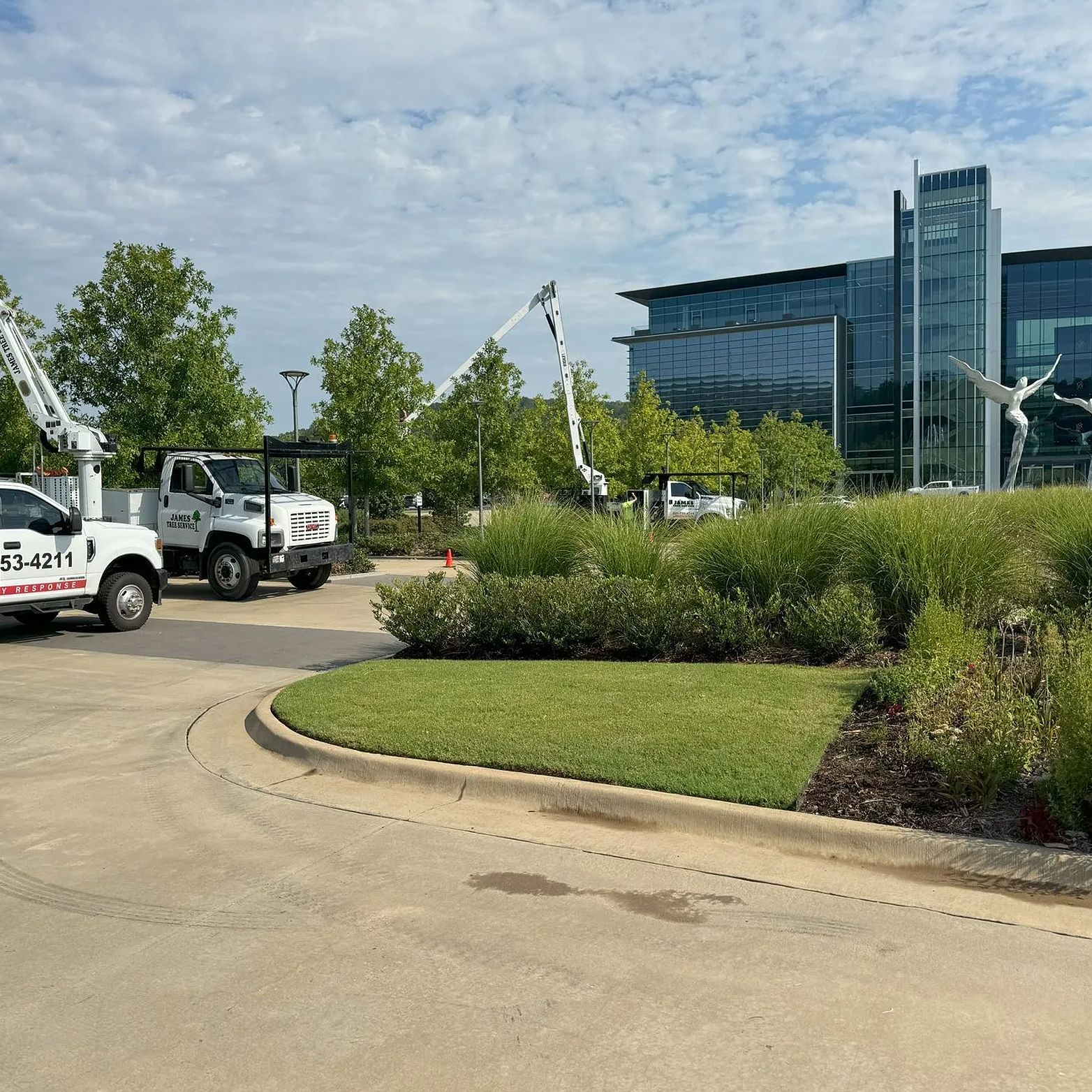 Three tree service trucks trimming trees in front of a modern building with sculptures.