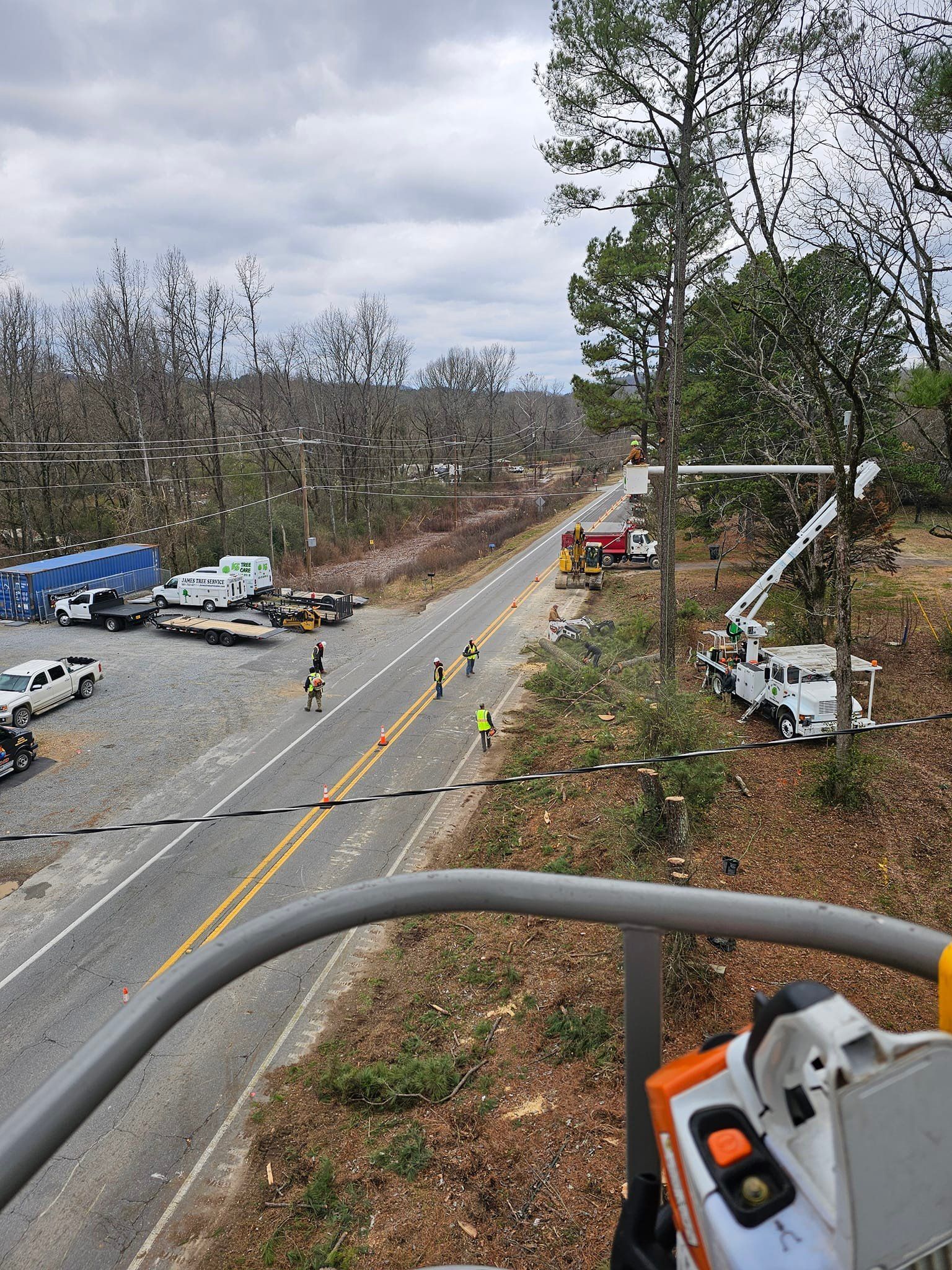 Utility trucks and workers on a road repairing power lines; chainsaw in foreground, cloudy sky.
