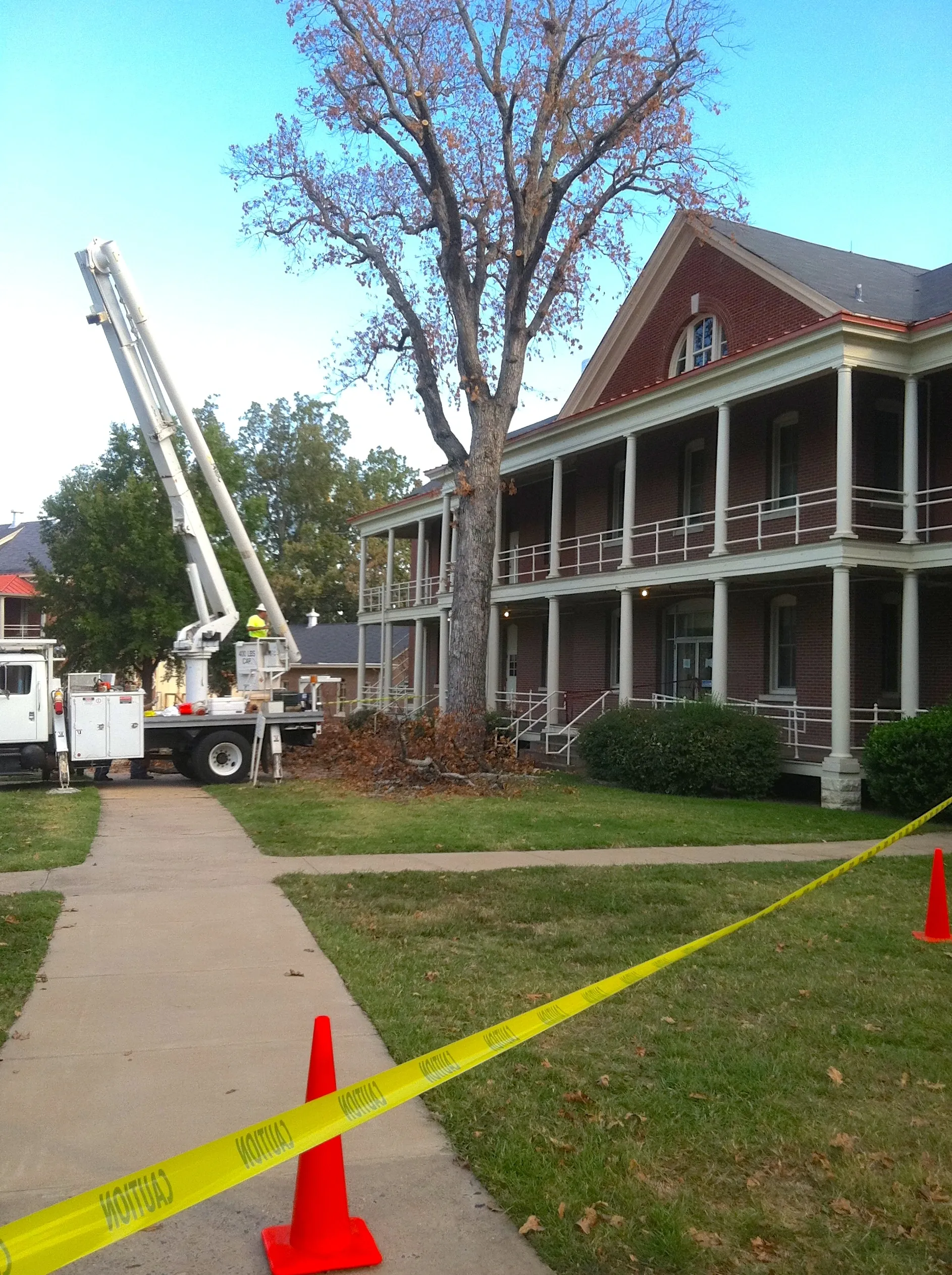 Tree trimming near a historic building. A worker on a truck lift trims branches. Caution tape.