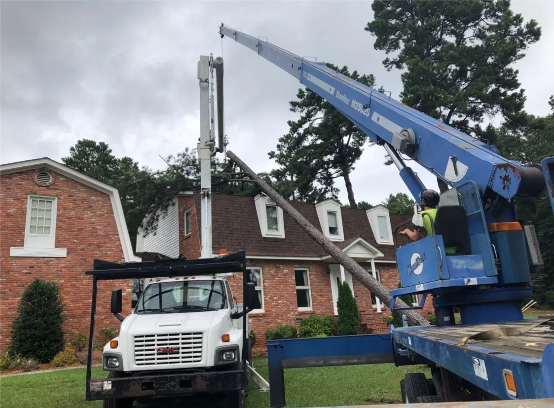 A utility truck with a crane lifting a telephone pole near a brick house.