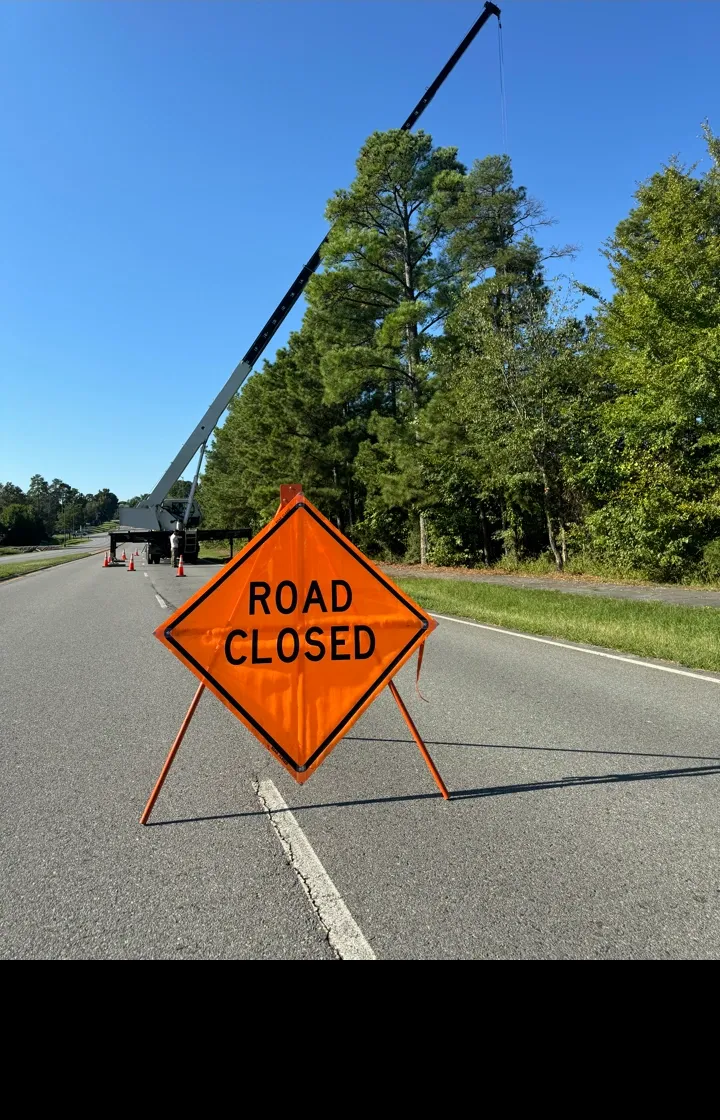 Road closed sign on an empty highway; a crane and trees are in the background under a blue sky.