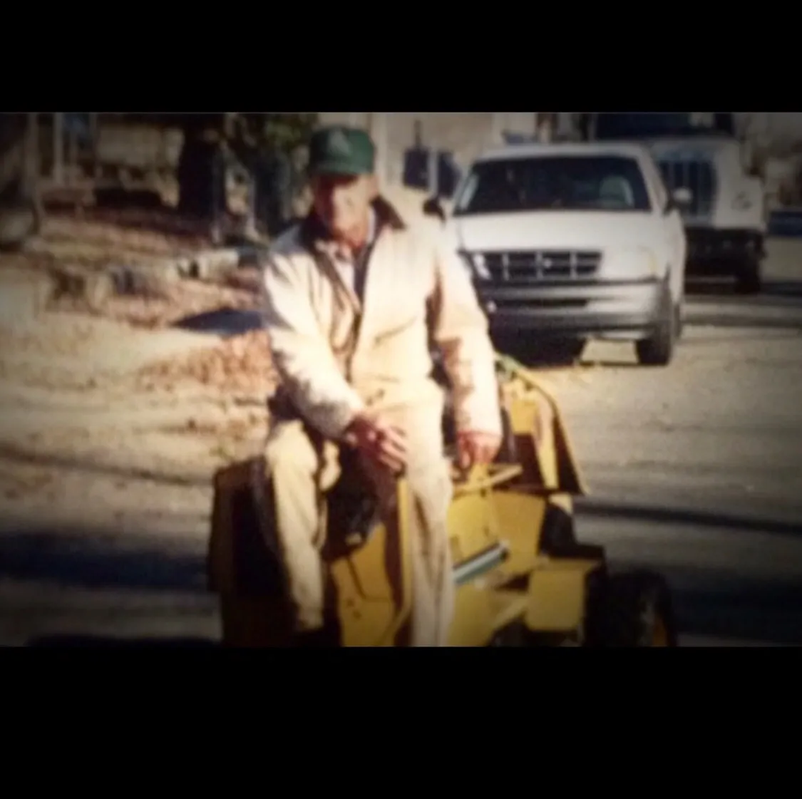 Man in tan jumpsuit sits on yellow lawnmower in street, with white truck behind him.