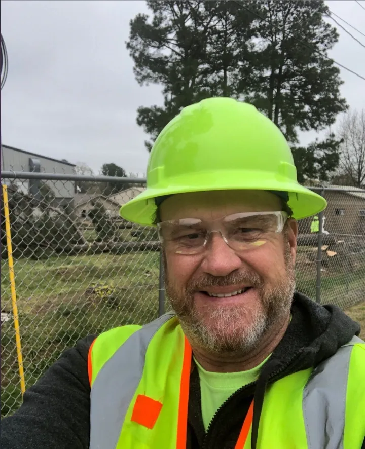 Man in neon green safety gear smiles outdoors near a fence and building.