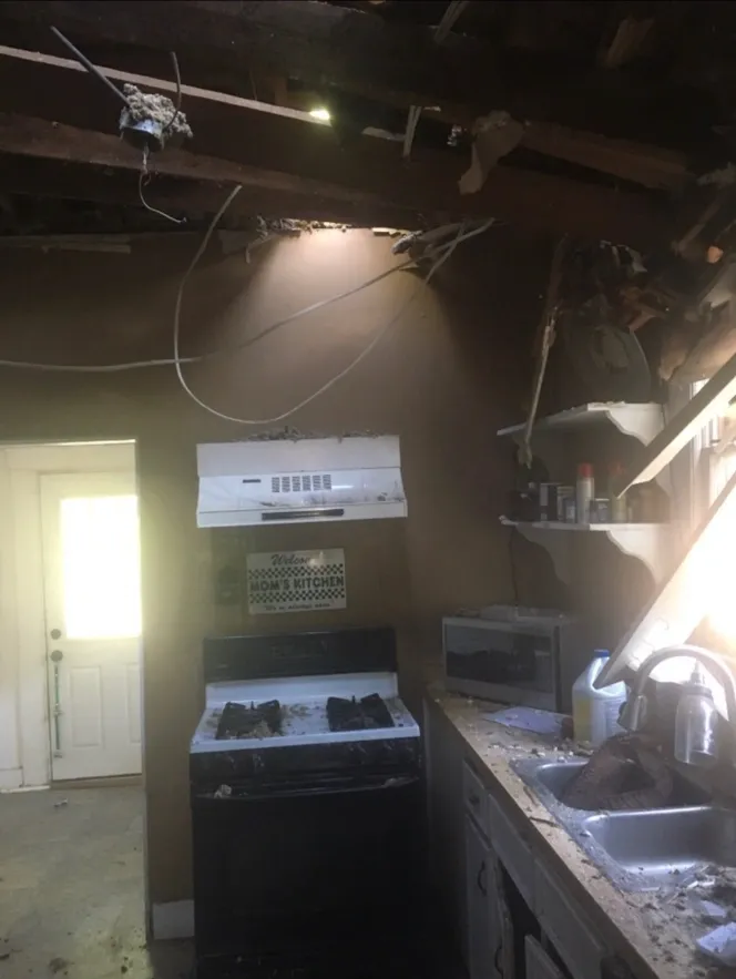 Interior view of a damaged kitchen, with a stove, sink, and exposed ceiling. Light streams in from above.