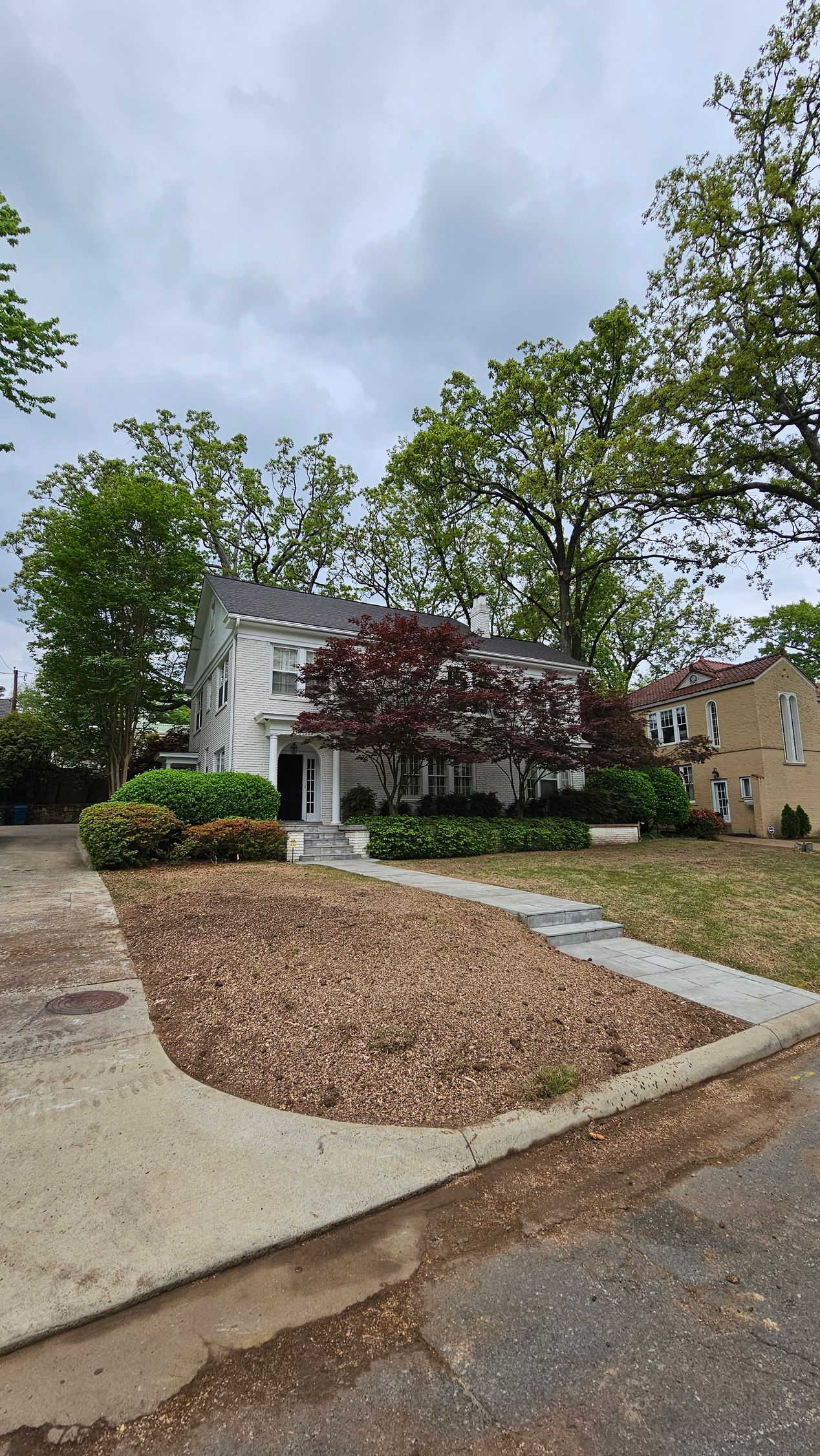 A two-story white house with a brown mulch yard and green trees under a cloudy sky.