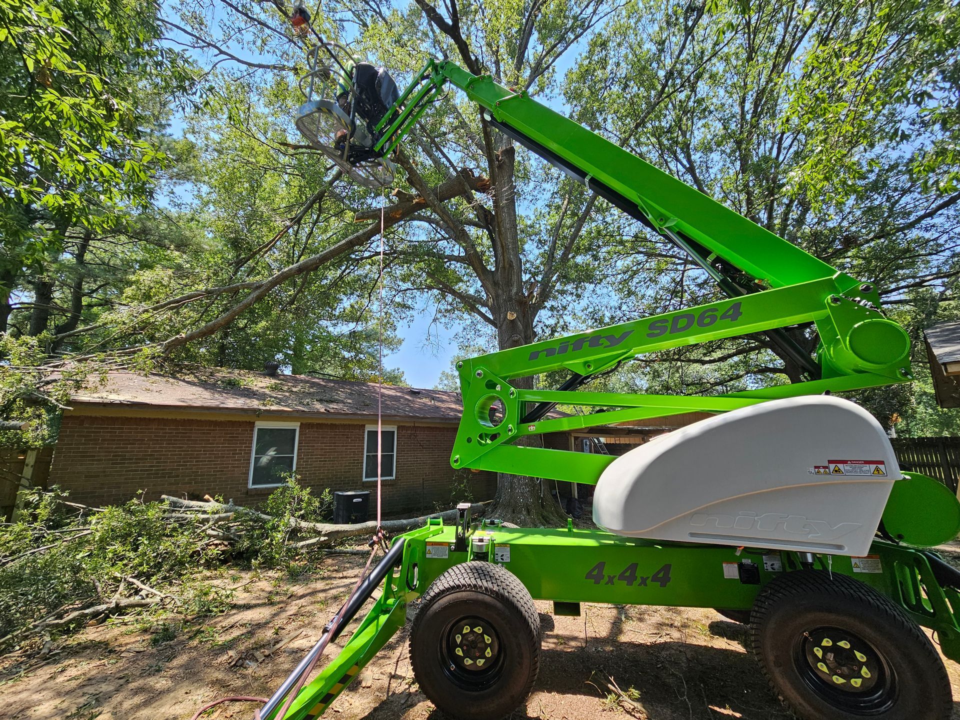 Green lift reaching toward tree branches; worker trimming tree near a house.
