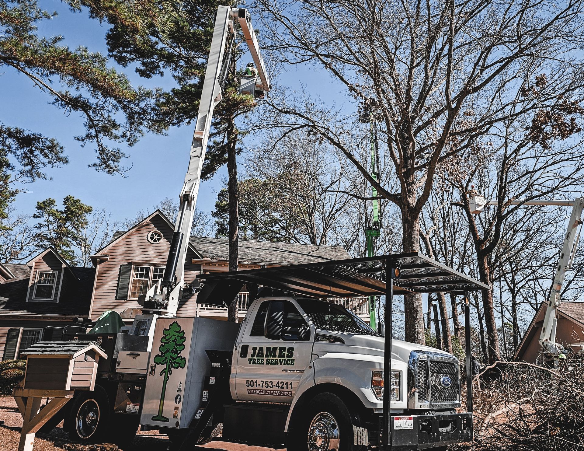 White tree service truck with extended boom trimming trees near a house under a clear blue sky.