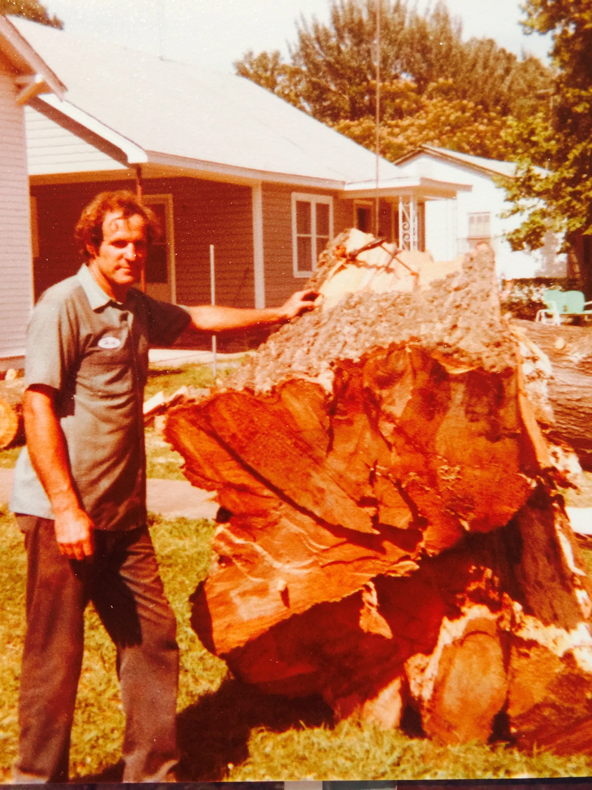 Man stands next to a large tree trunk, outdoors. He is wearing a gray shirt and dark pants.