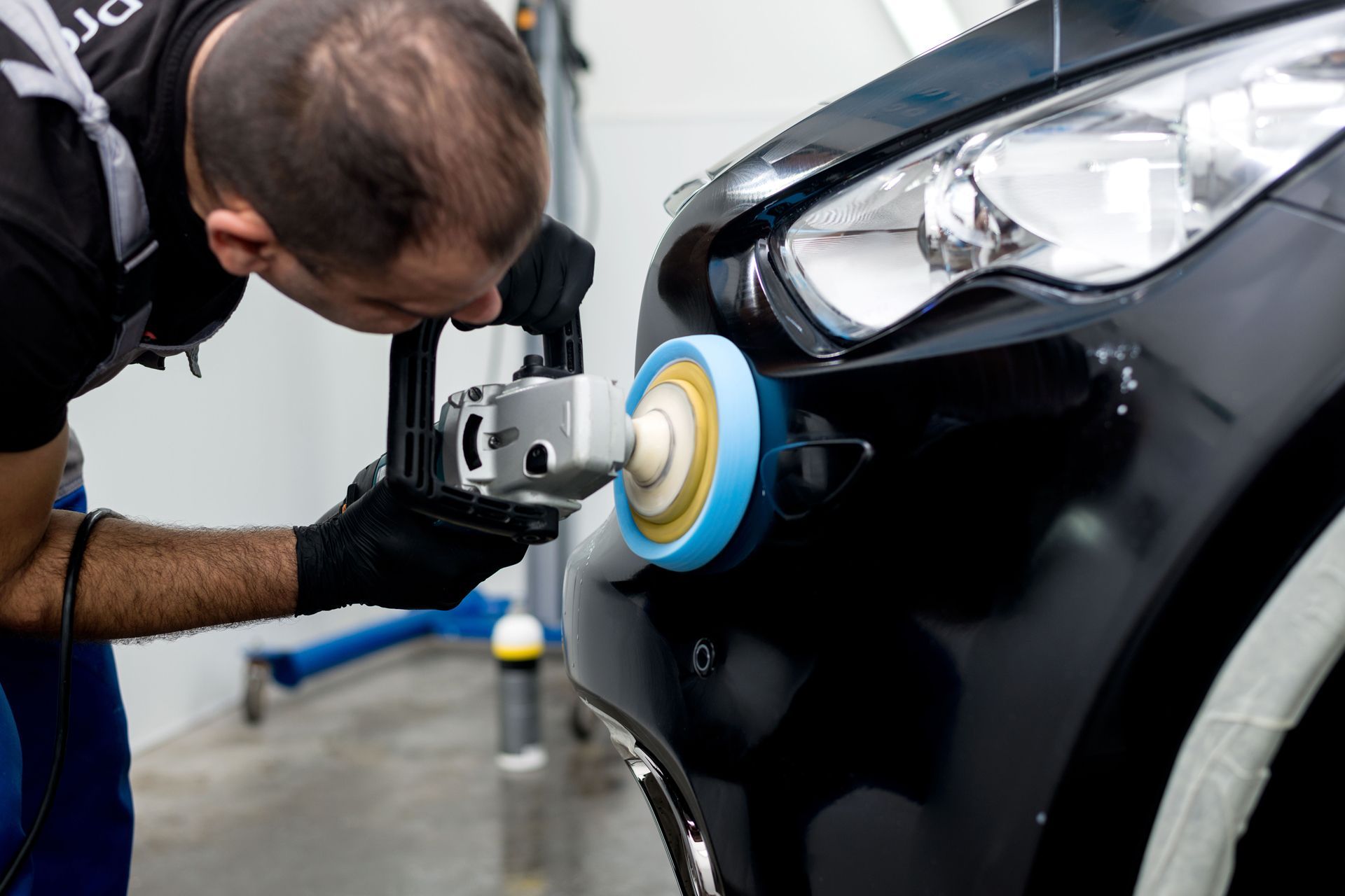Person polishing black car bumper with power tool in a garage.