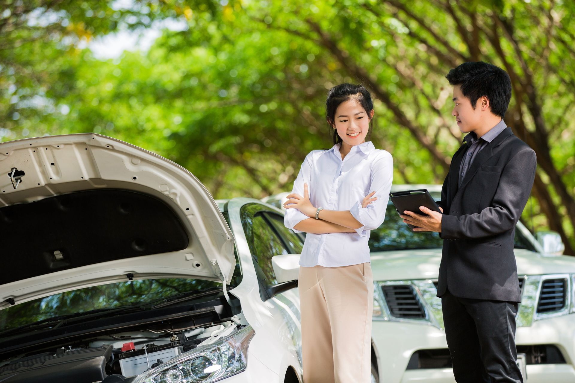 Woman and car salesman near two white cars with hoods open, discussing with a tablet outdoors.