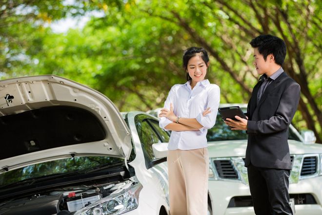 Woman and car salesman near two white cars with hoods open, discussing with a tablet outdoors.