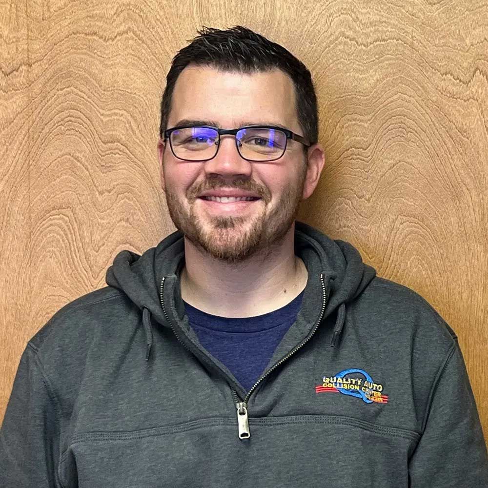 Man with glasses and a beard smiling, wearing a gray hooded sweatshirt, standing in front of a wood background.