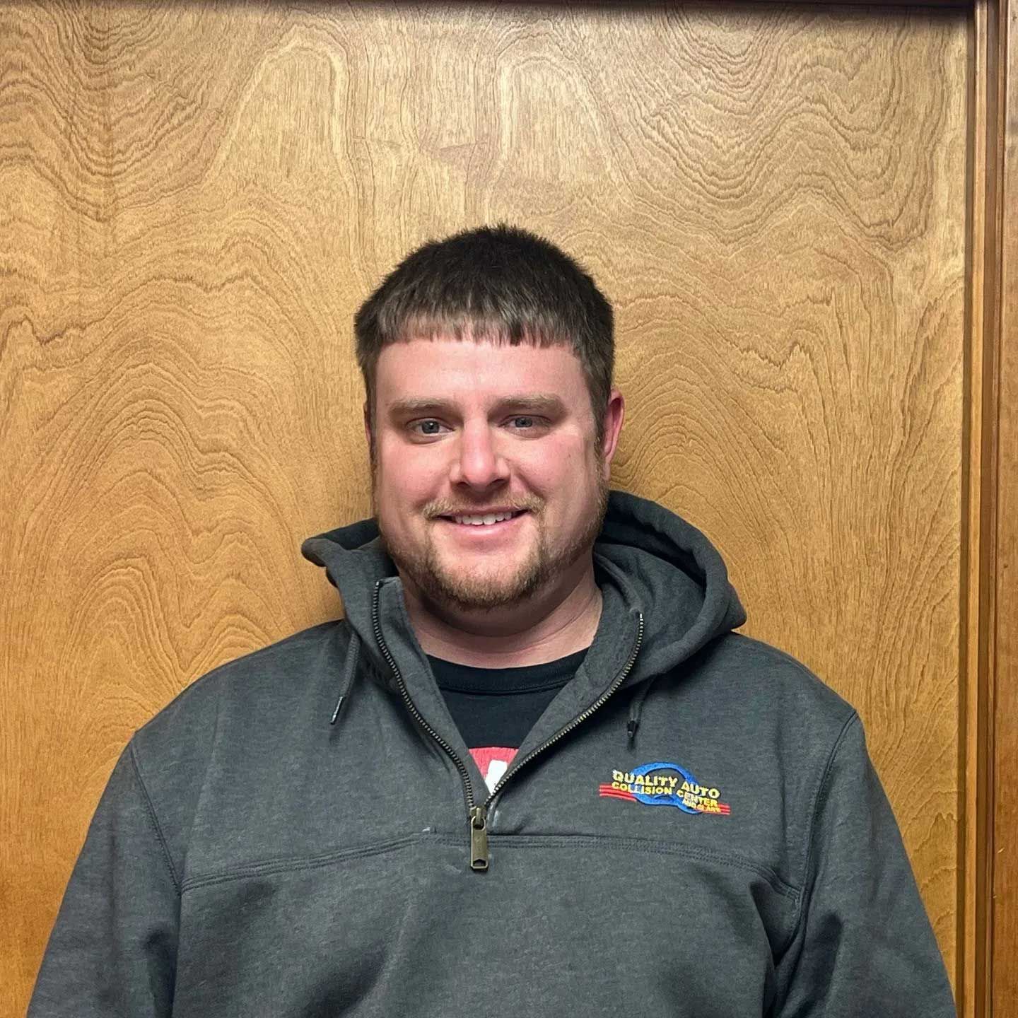 Man smiling, wearing a gray hooded jacket, standing in front of a wood door.