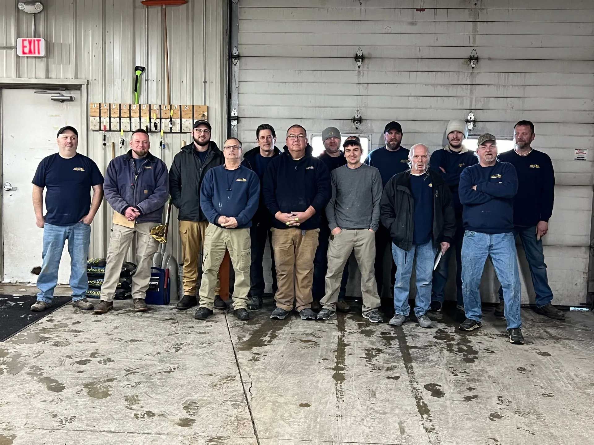 Group of men in a garage. Most wear navy shirts and jeans, posing near a garage door and interior door.