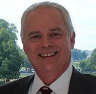 Smiling man in a suit and tie outdoors with trees and water in the background