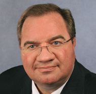 Headshot of a man wearing glasses and a dark suit against a gray background