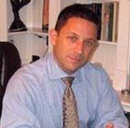 Man seated indoors, wearing a light blue shirt and patterned tie, looking at the camera.