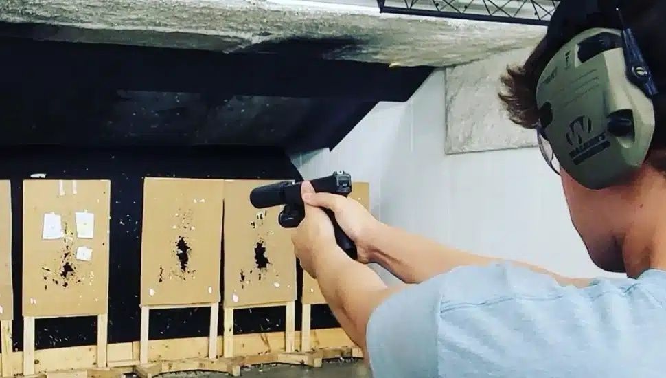 Person aiming a handgun at paper targets at an indoor shooting range, wearing ear protection.