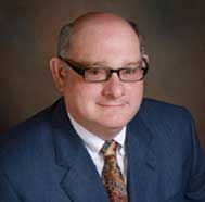 Portrait of a man in a blue suit and patterned tie, wearing glasses, against a gray background