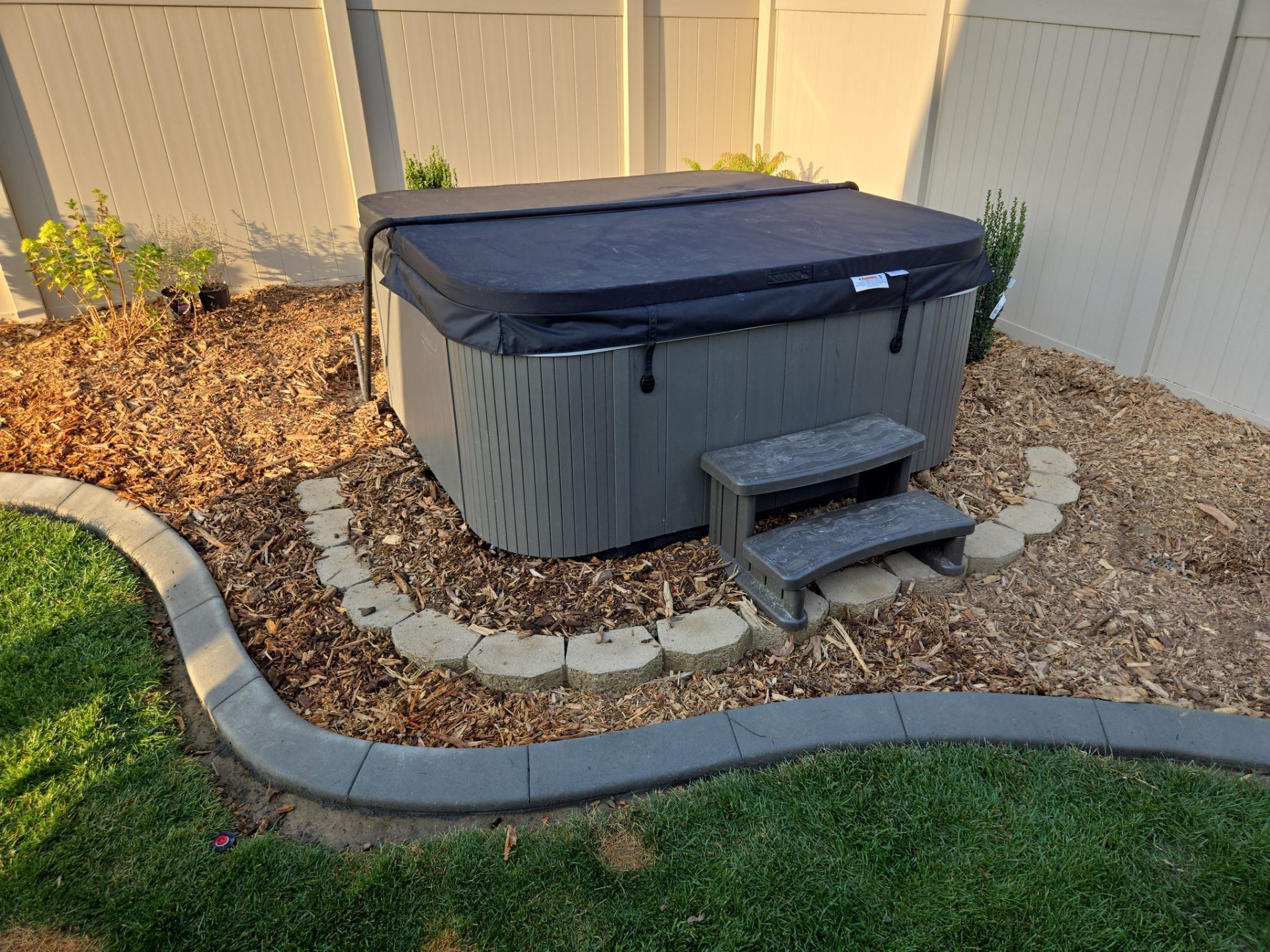 Hot tub with cover and steps in a backyard, surrounded by mulch and edging.