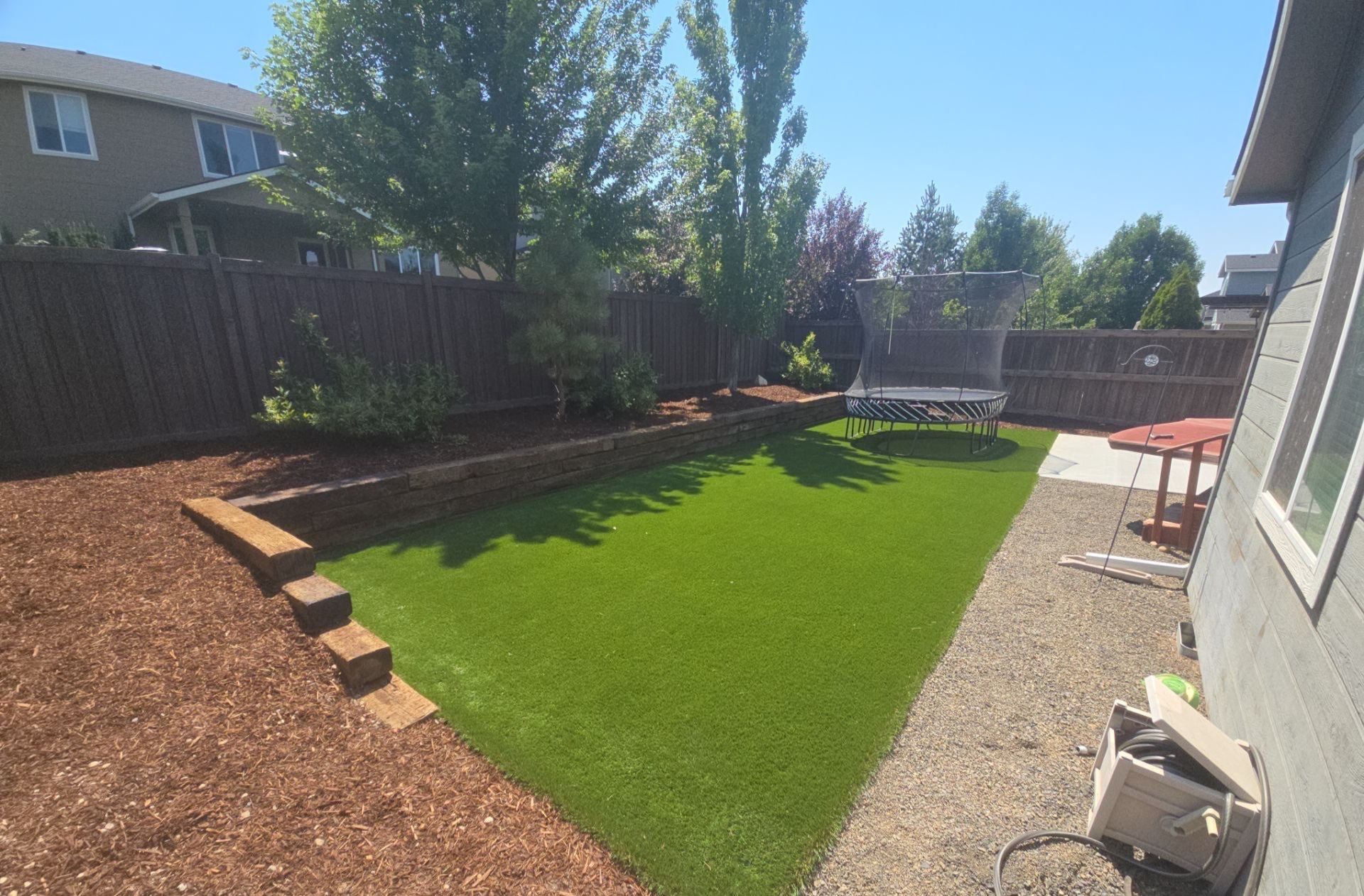 Backyard with artificial turf, surrounded by a wooden fence and mulch. A trampoline sits on the grass.