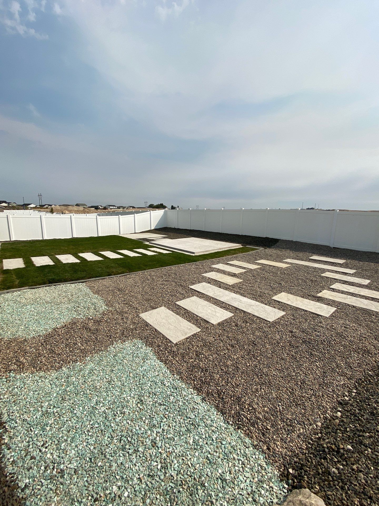 Backyard with stone walkways, grass, and gravel. White walls and cloudy sky.