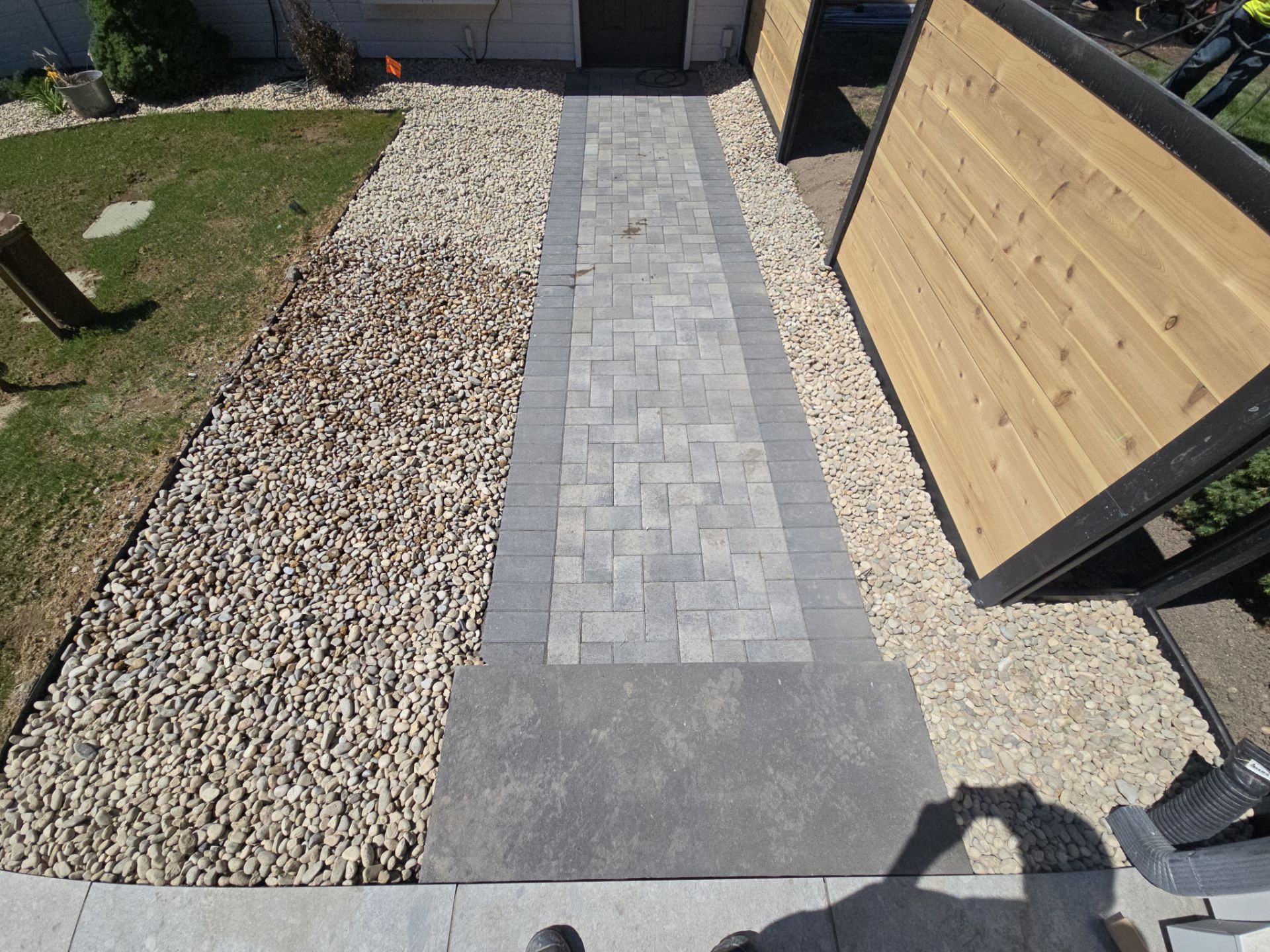A pathway of pavers leads to a building entrance, flanked by gravel and wooden fencing, with a green lawn.