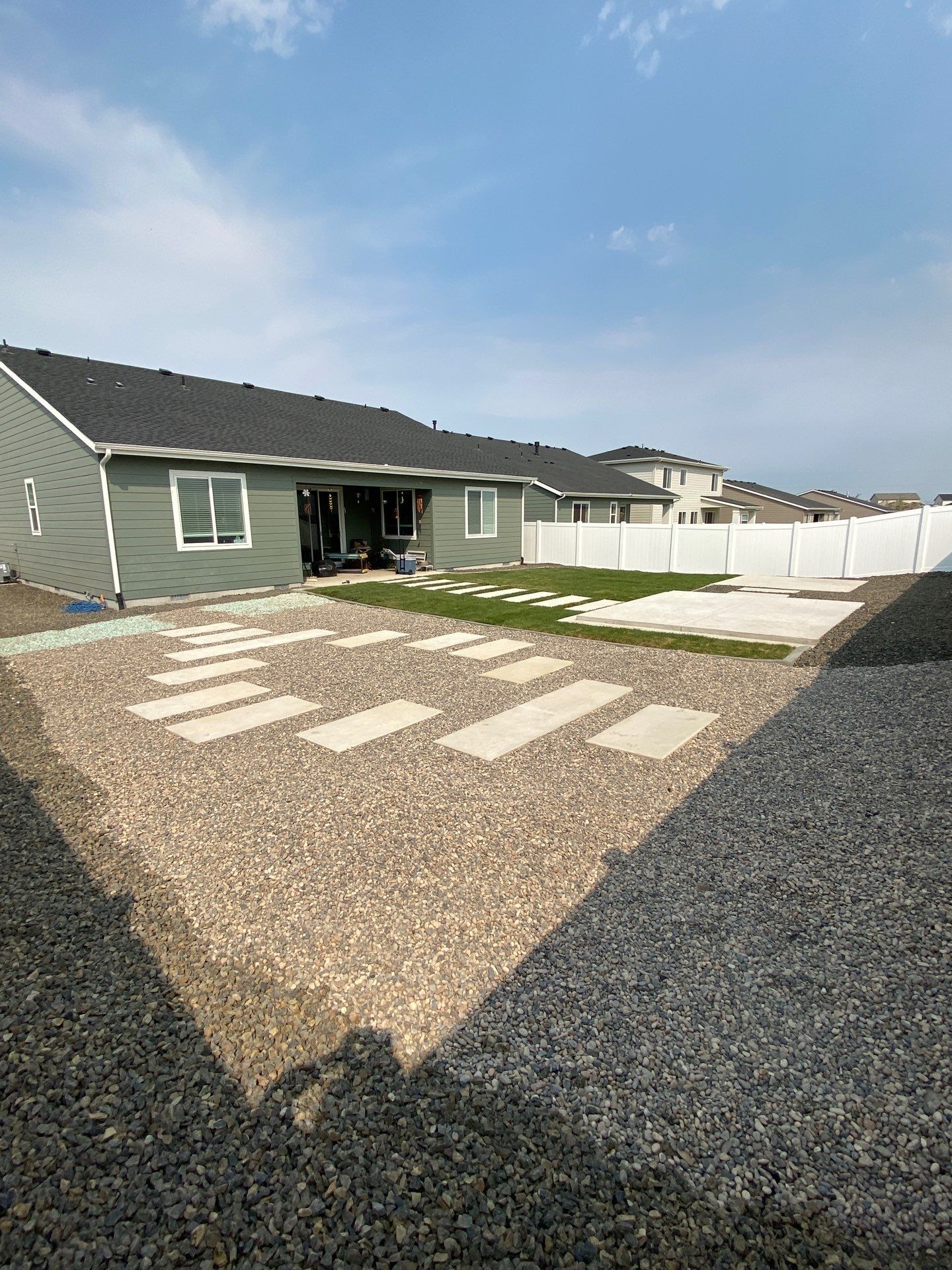Backyard with stepping stones on gravel, light green house, white fence. Sunny day.