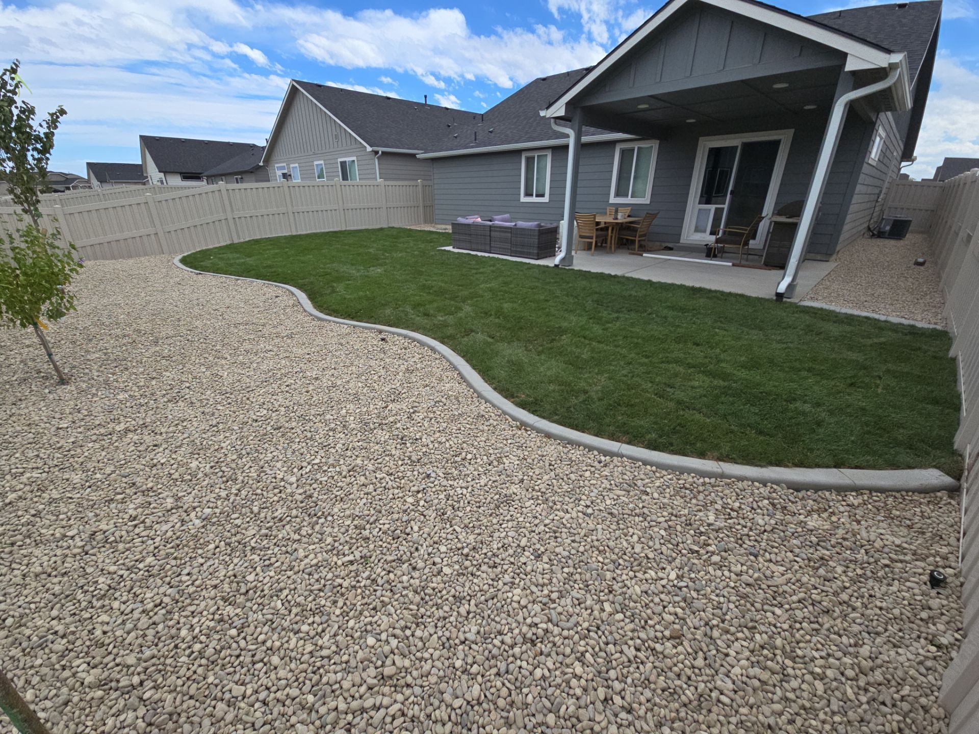 Backyard with a green lawn bordered by tan gravel and a house with a covered patio.