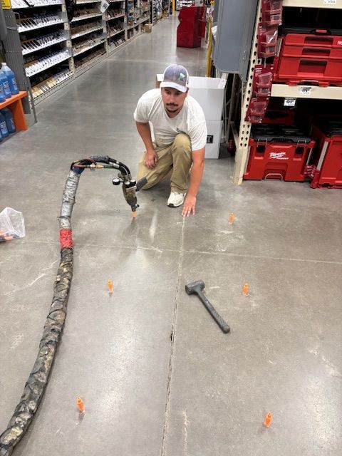 Man kneels to work on a floor crack in a hardware store. He points at the crack and the tools around it.