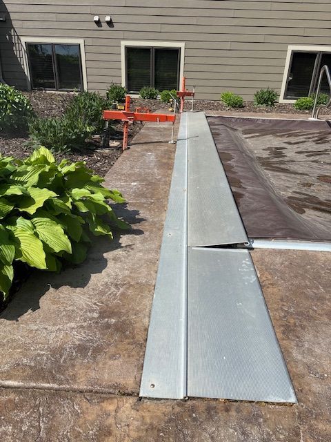 Poolside walkway with metal edge, pool cover, orange tool, and bushes.