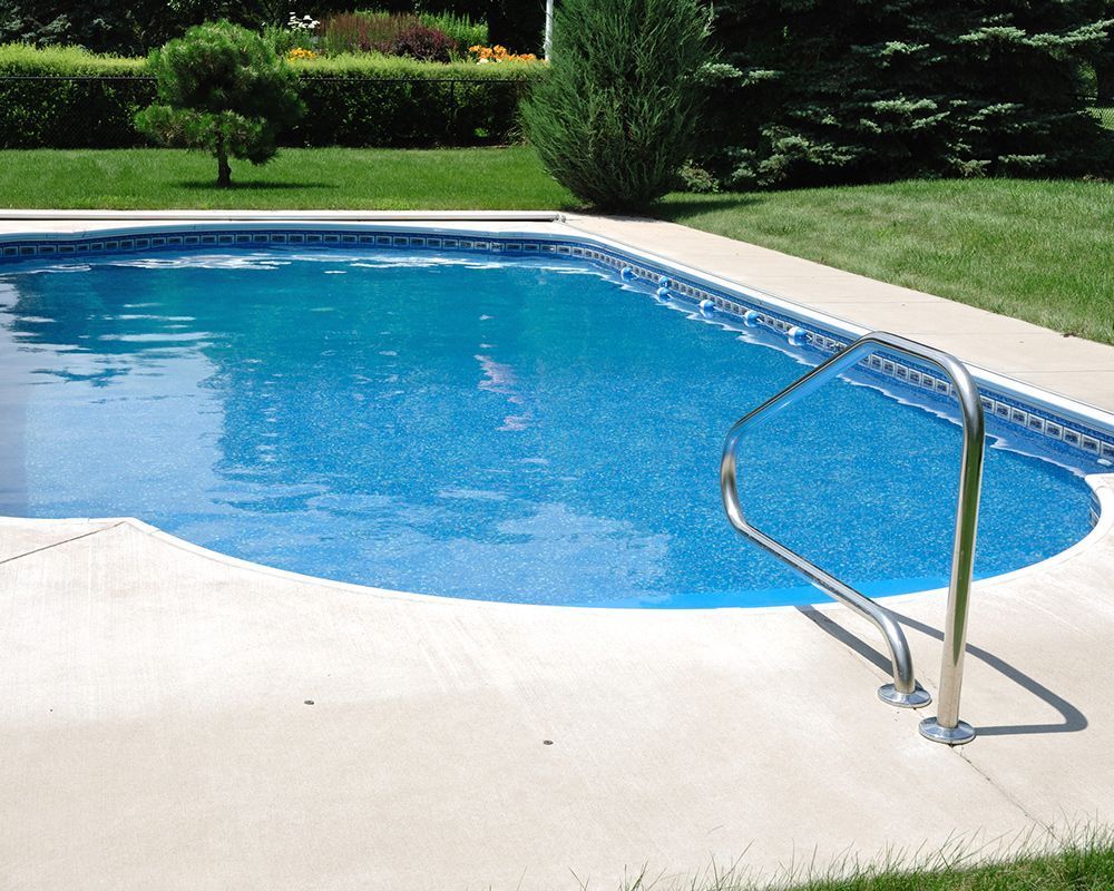 Swimming pool with concrete deck and stainless steel handrail, surrounded by green grass and shrubs.
