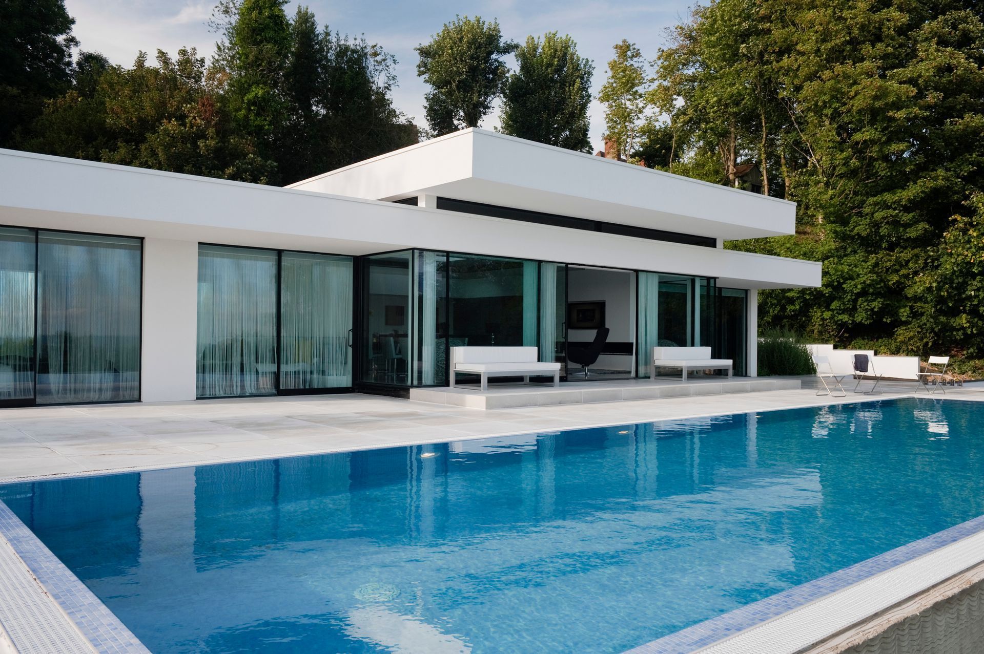 Modern white house with a pool, sliding glass doors, and a backdrop of trees.